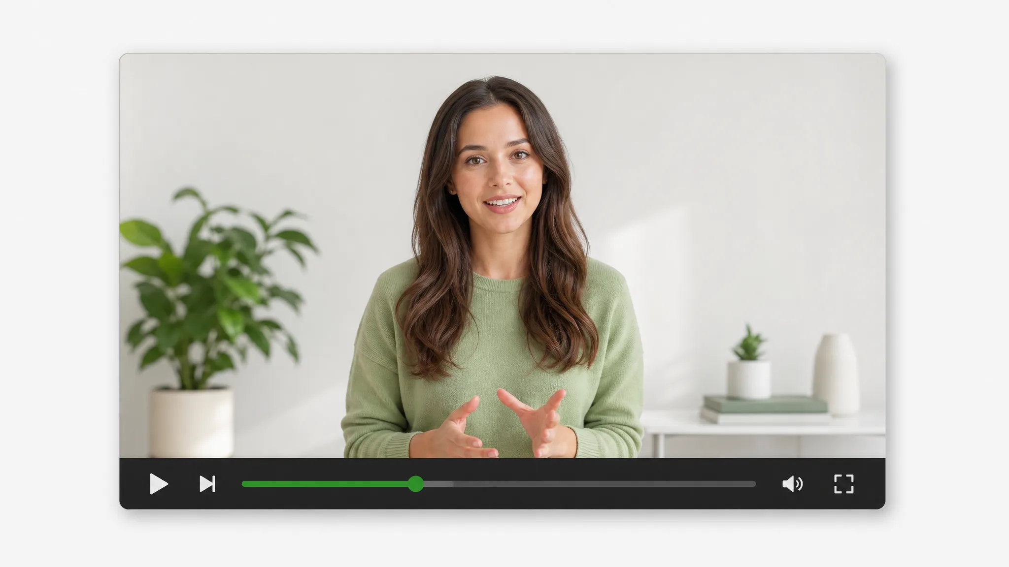 A woman wearing a green sweater talks in a bright room beside a plant, shown in a video player with play and volume controls.